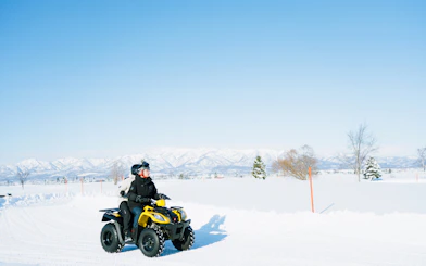 Two people riding an ATV on a snowy landscape in Bibai, Hokkaido, with mountains in the background.