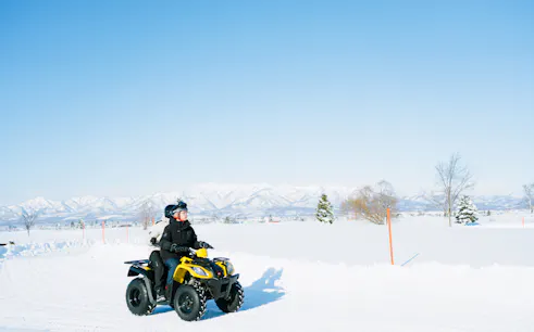 Two people riding an ATV on a snowy landscape in Bibai, Hokkaido, with mountains in the background.