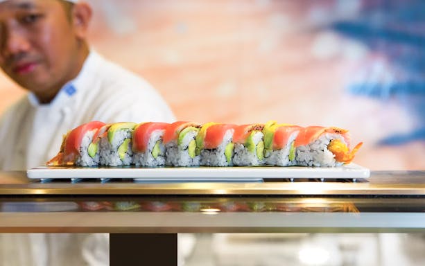 Sushi chef presenting a colorful sushi roll on a white plate.