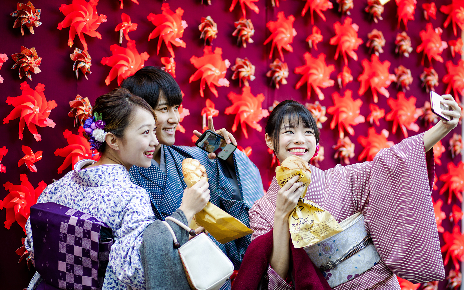 Friends in kimonos taking a selfie with red pinwheel backdrop in Japan.