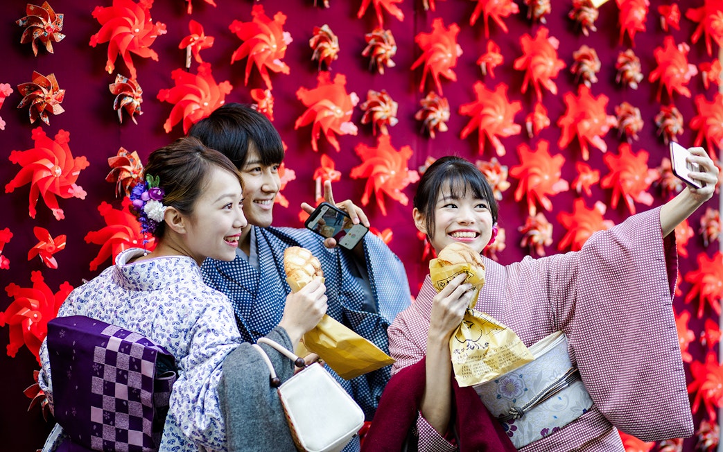 Friends in kimonos taking a selfie with red pinwheel backdrop in Japan.