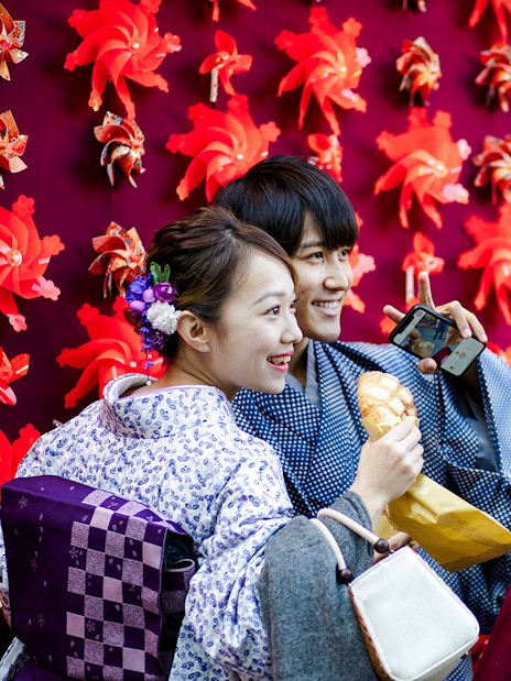 Friends in kimonos taking a selfie with red pinwheel backdrop in Japan.