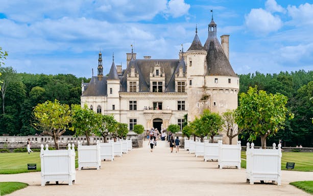 Chenonceau Castle exterior with visitors walking along the tree-lined path.