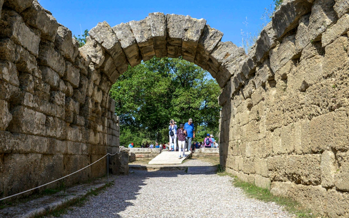Visitors walking through ancient stone archway at Olympia, Greece.