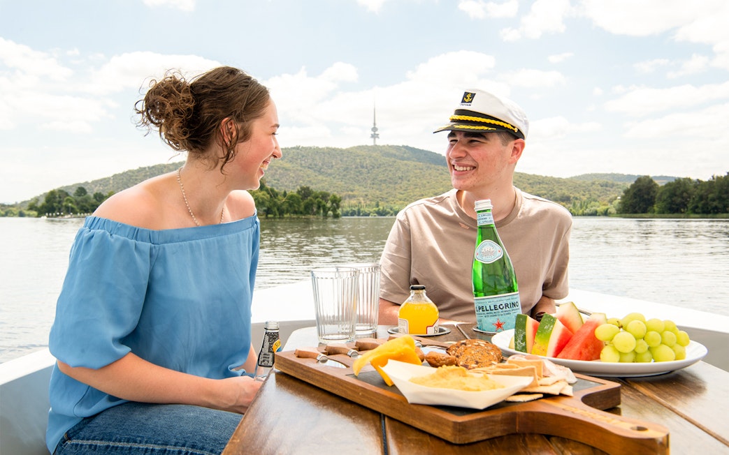 Couple enjoying a picnic on an electric boat with views of Black Mountain Tower, Canberra.