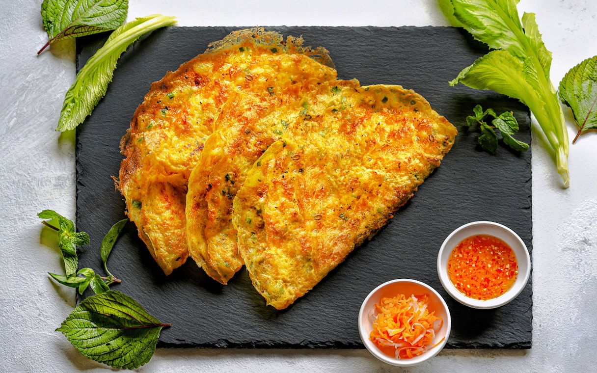 Banh Xeo Vietnamese crepe pancake with herbs and dipping sauce on slate plate.