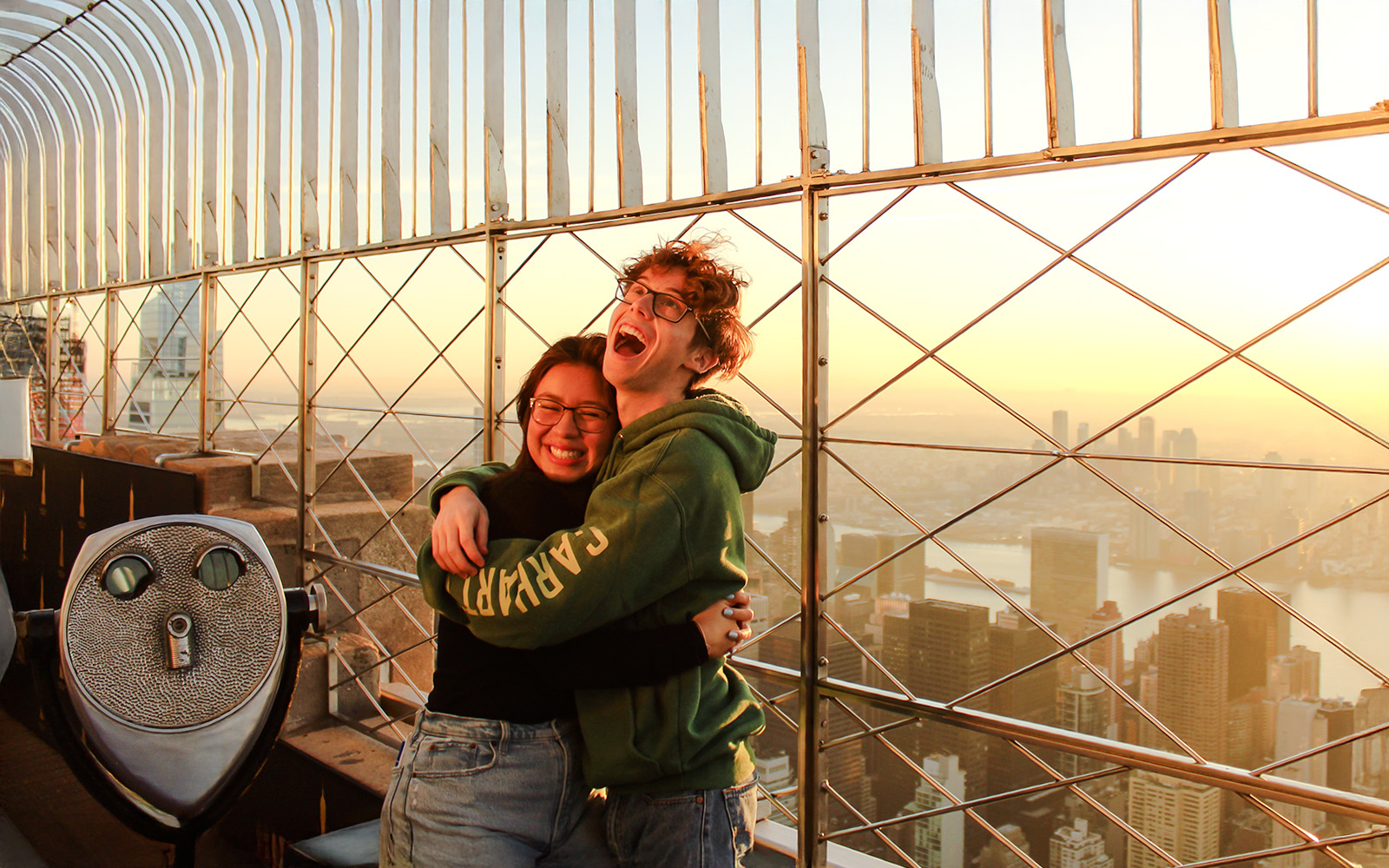 Guests embracing at Empire State Building observatory with New York City skyline at sunset.