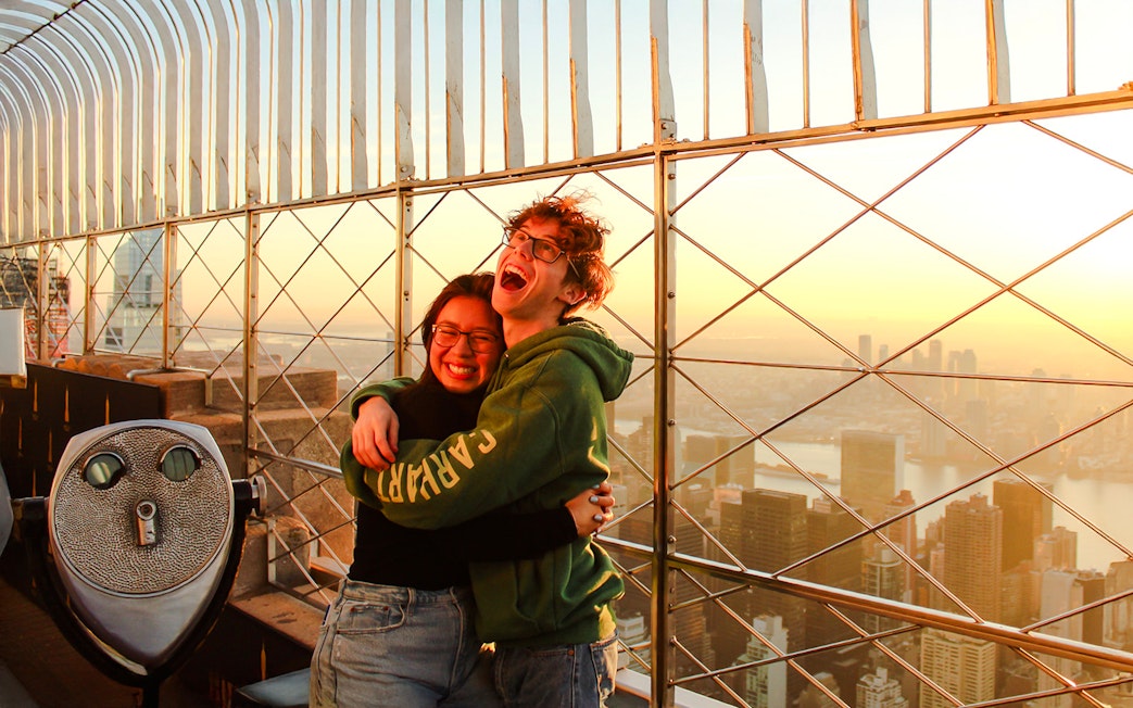 Guests embracing at Empire State Building observatory with New York City skyline at sunset.
