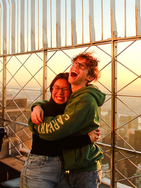 Guests embracing at Empire State Building observatory with New York City skyline at sunset.