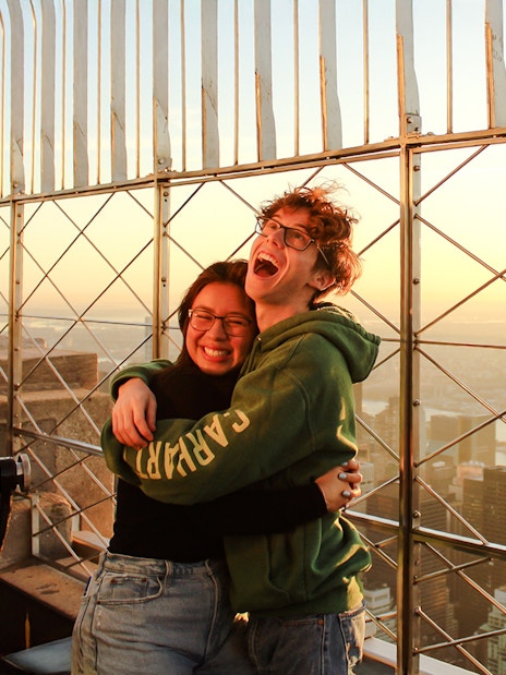 Guests embracing at Empire State Building observatory with New York City skyline at sunset.