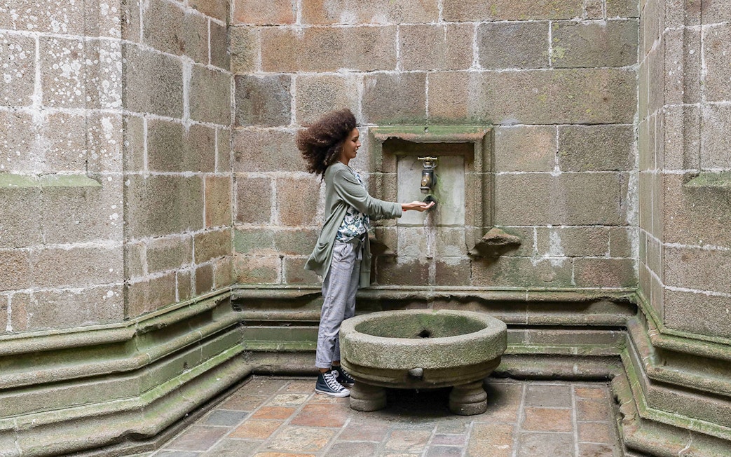 Tourist at Mont-Saint-Michel Abbey using historic water fountain in Normandy, France.