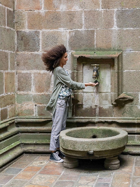 Tourist at Mont-Saint-Michel Abbey using historic water fountain in Normandy, France.