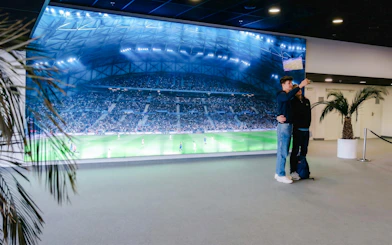 Visitors taking a selfie in front of a large screen displaying a soccer match at OM Stadium.