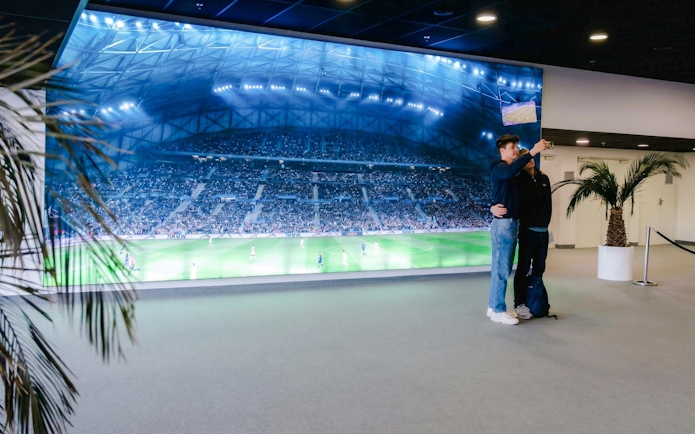 Visitors taking a selfie in front of a large screen displaying a soccer match at OM Stadium.