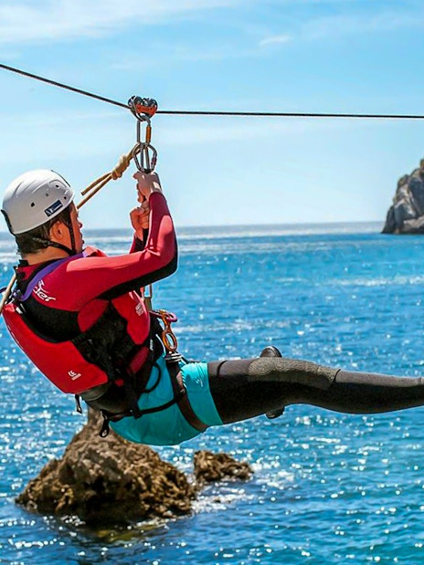 Person ziplining over ocean during coasteering adventure near rocky cliffs.