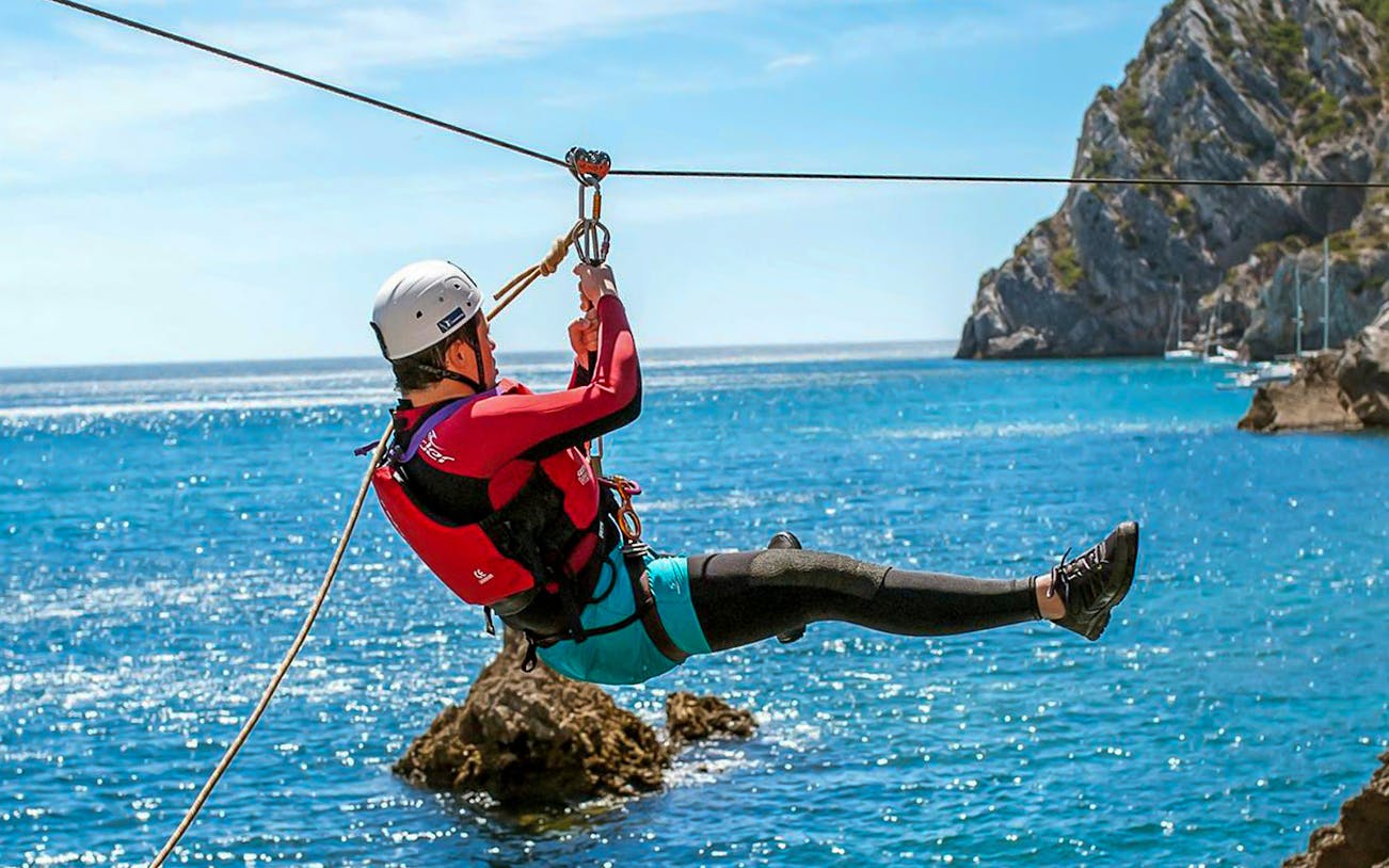 Person ziplining over ocean during coasteering adventure near rocky cliffs.