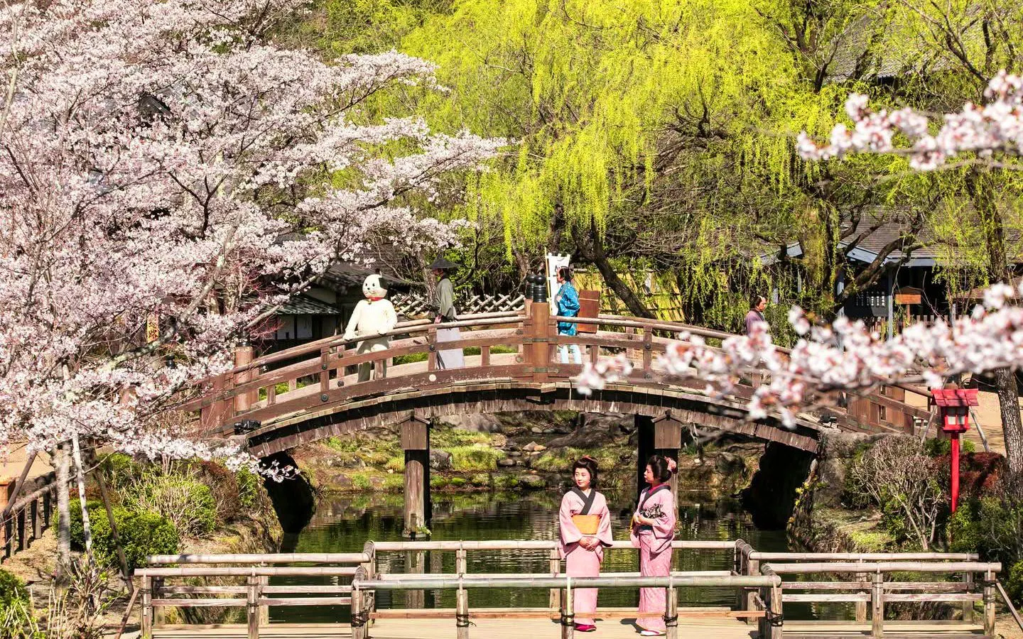 Visitors in traditional attire at Edo Wonderland Nikko Edomura, Japan, with cherry blossoms and a wooden bridge.