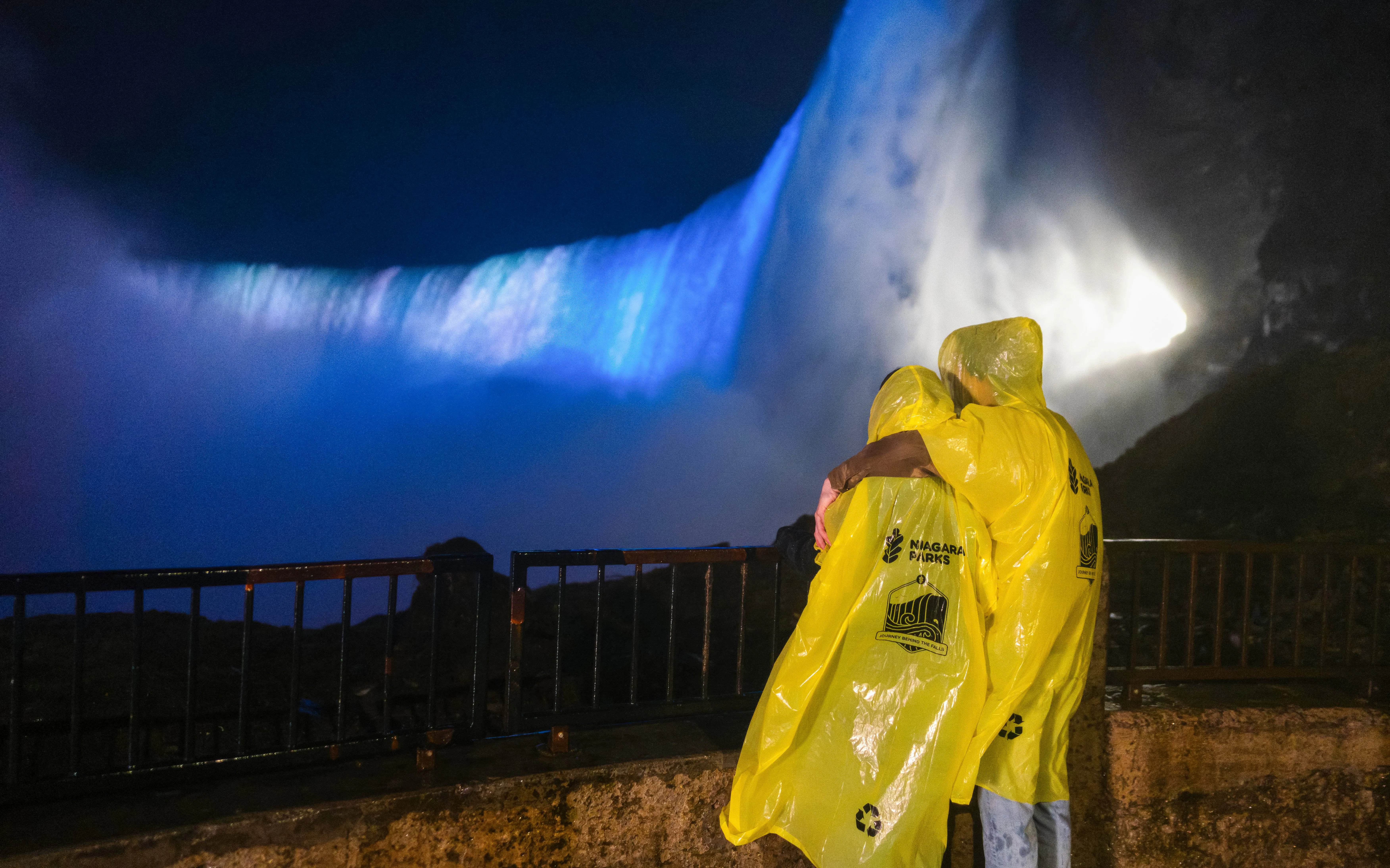 Visitors in yellow ponchos view illuminated Niagara Falls at night from a railing.
