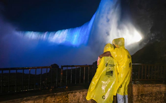 Visitors in yellow ponchos view illuminated Niagara Falls at night from a railing.