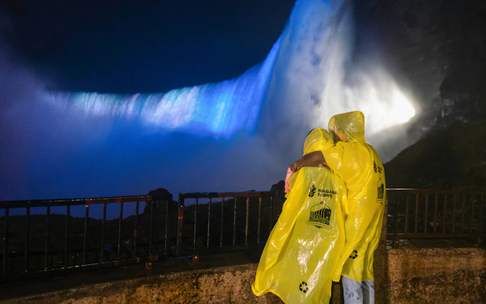 Visitors in yellow ponchos view illuminated Niagara Falls at night from a railing.
