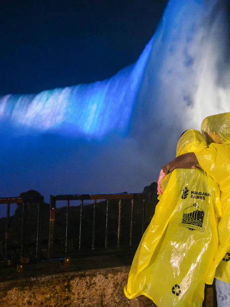 Visitors in yellow ponchos view illuminated Niagara Falls at night from a railing.