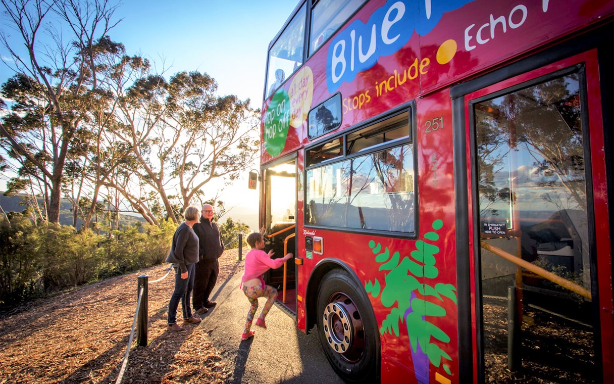 Tourists boarding a red double-decker bus in Sydney with scenic views in the background.