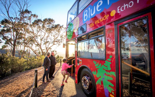 Tourists boarding a red double-decker bus in Sydney with scenic views in the background.