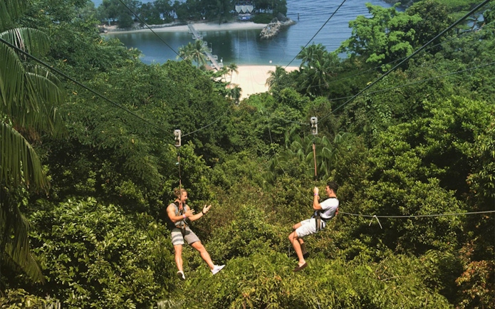 Two people ziplining over lush greenery towards a beach in Singapore.