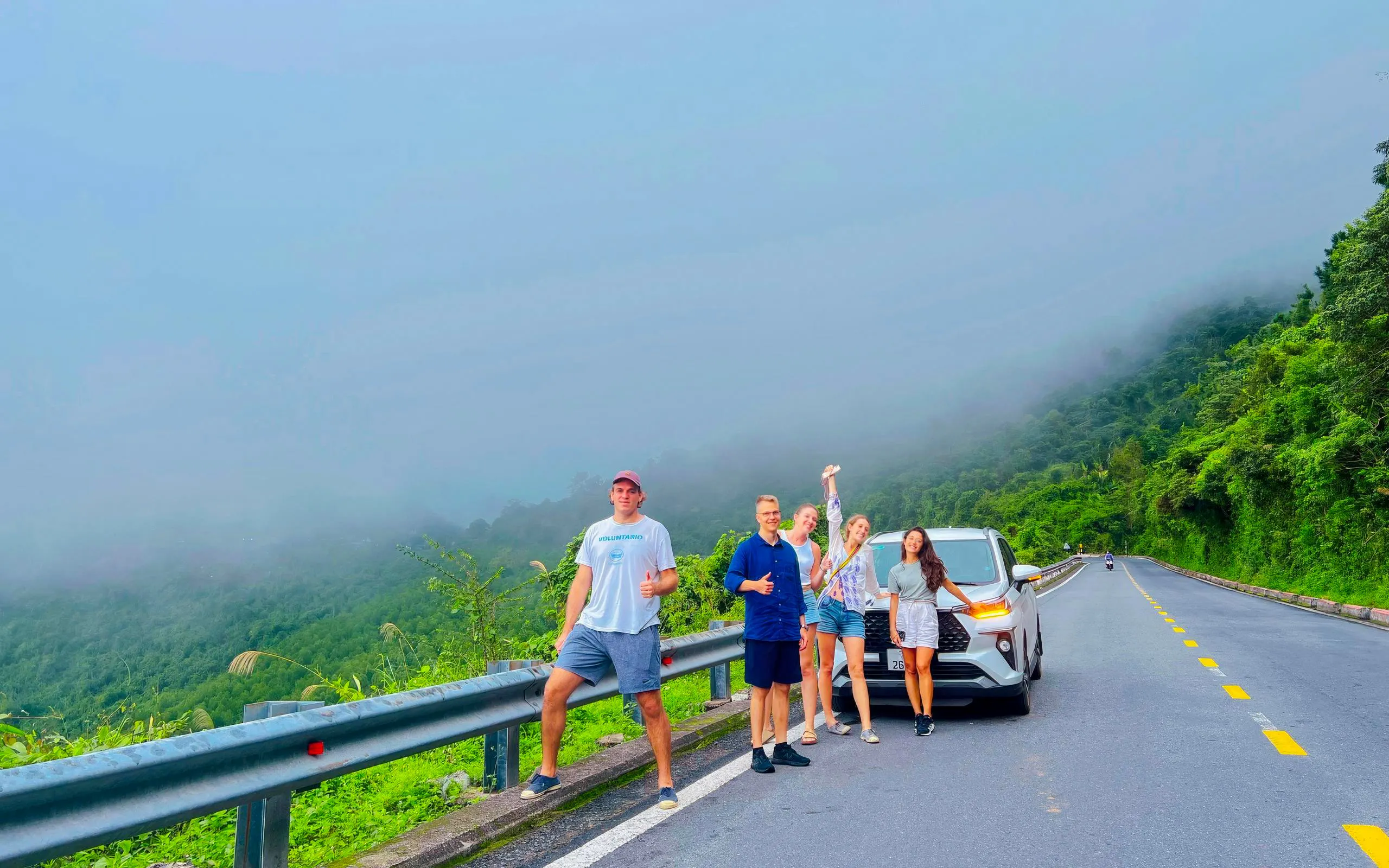 Group of tourists by car on Hai Van Pass road, lush greenery and misty hills in background, Vietnam.