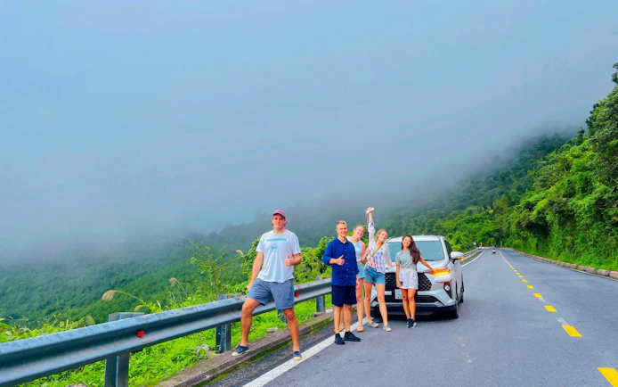 Group of tourists by car on Hai Van Pass road, lush greenery and misty hills in background, Vietnam.