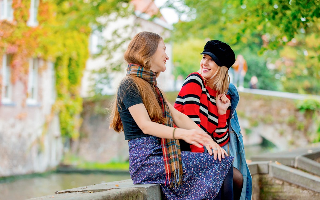 Two people sitting on a canal bridge in Bruges, surrounded by greenery.