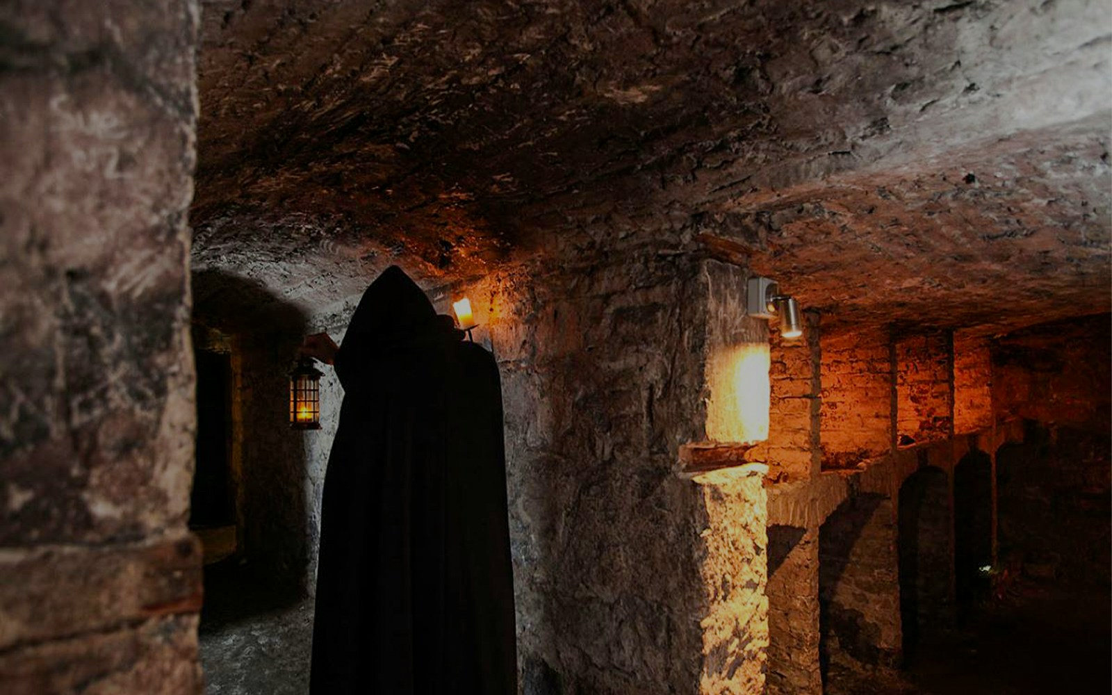 Cloaked figure holding lantern in Edinburgh underground vaults during haunted night tour.