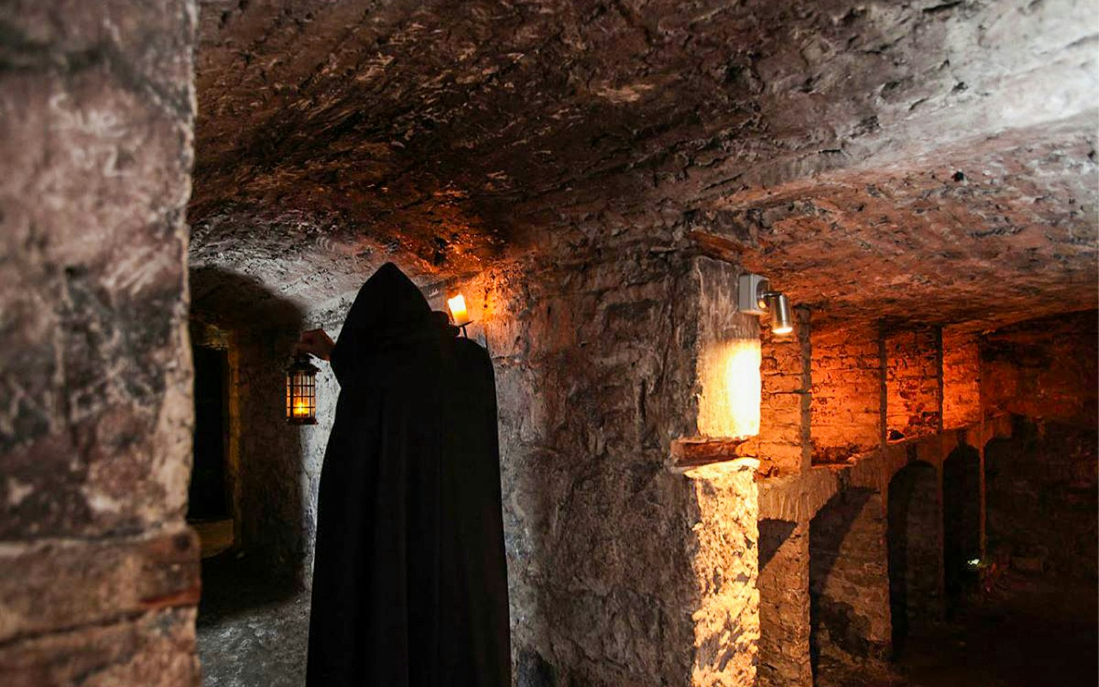 Cloaked figure holding lantern in Edinburgh underground vaults during haunted night tour.