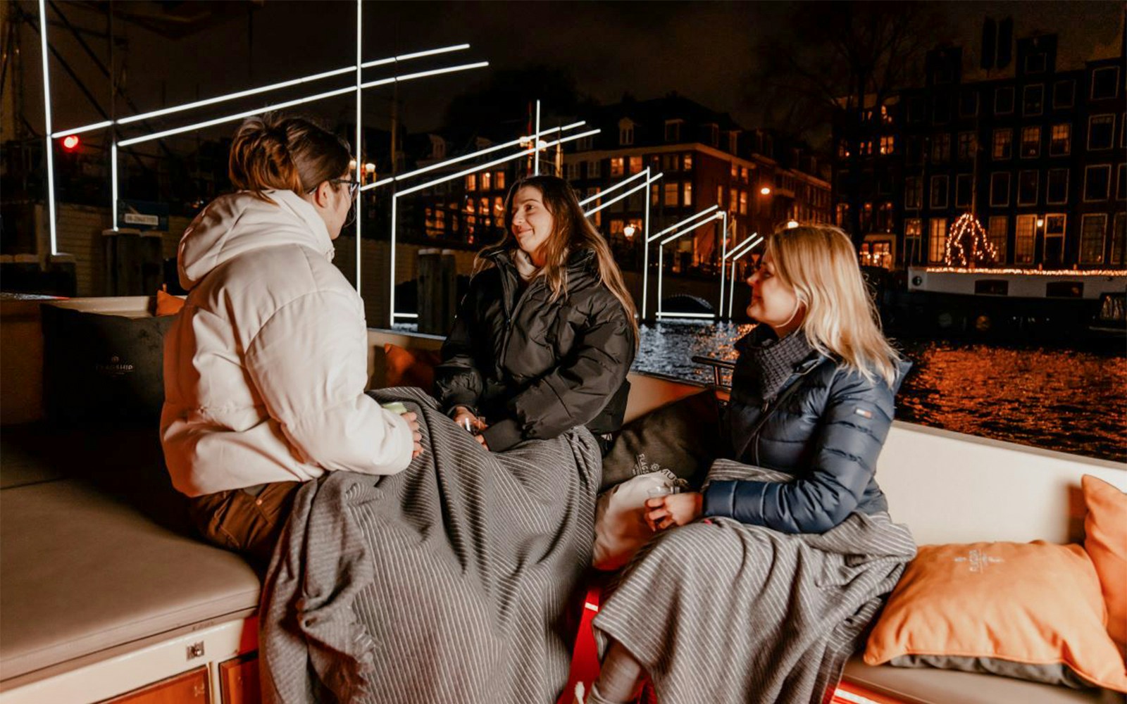 People enjoying Amsterdam Light Festival cruise with blankets on a canal boat at night.