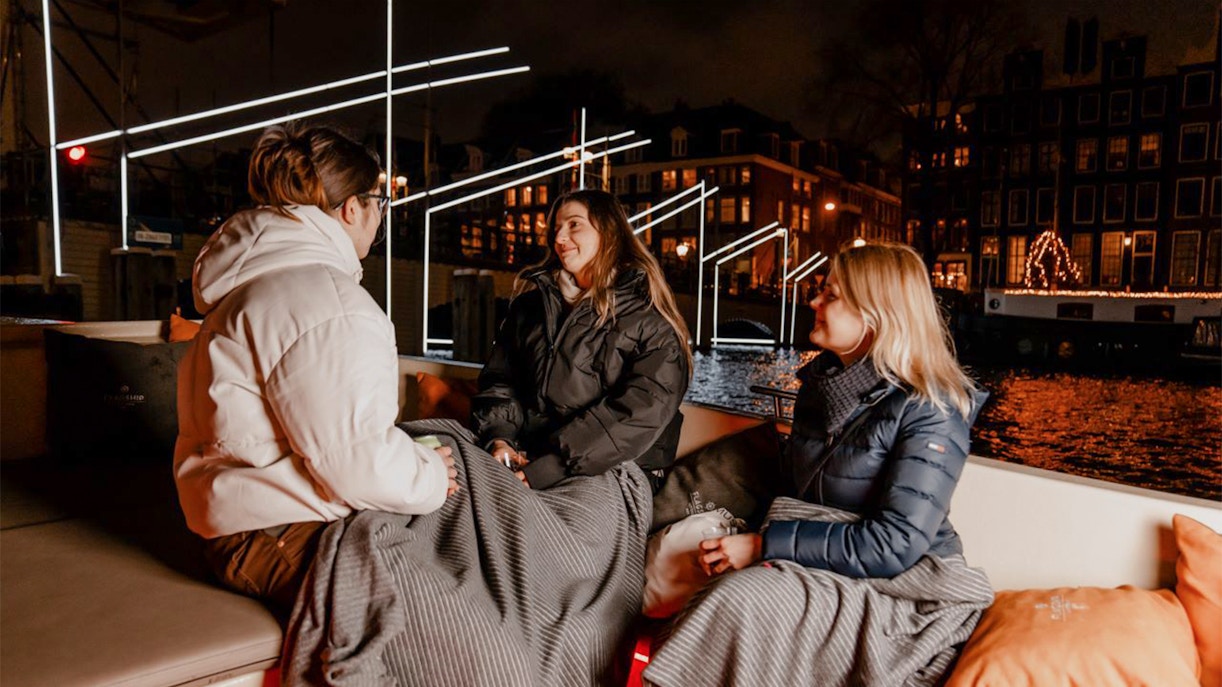 People enjoying Amsterdam Light Festival cruise with blankets on a canal boat at night.