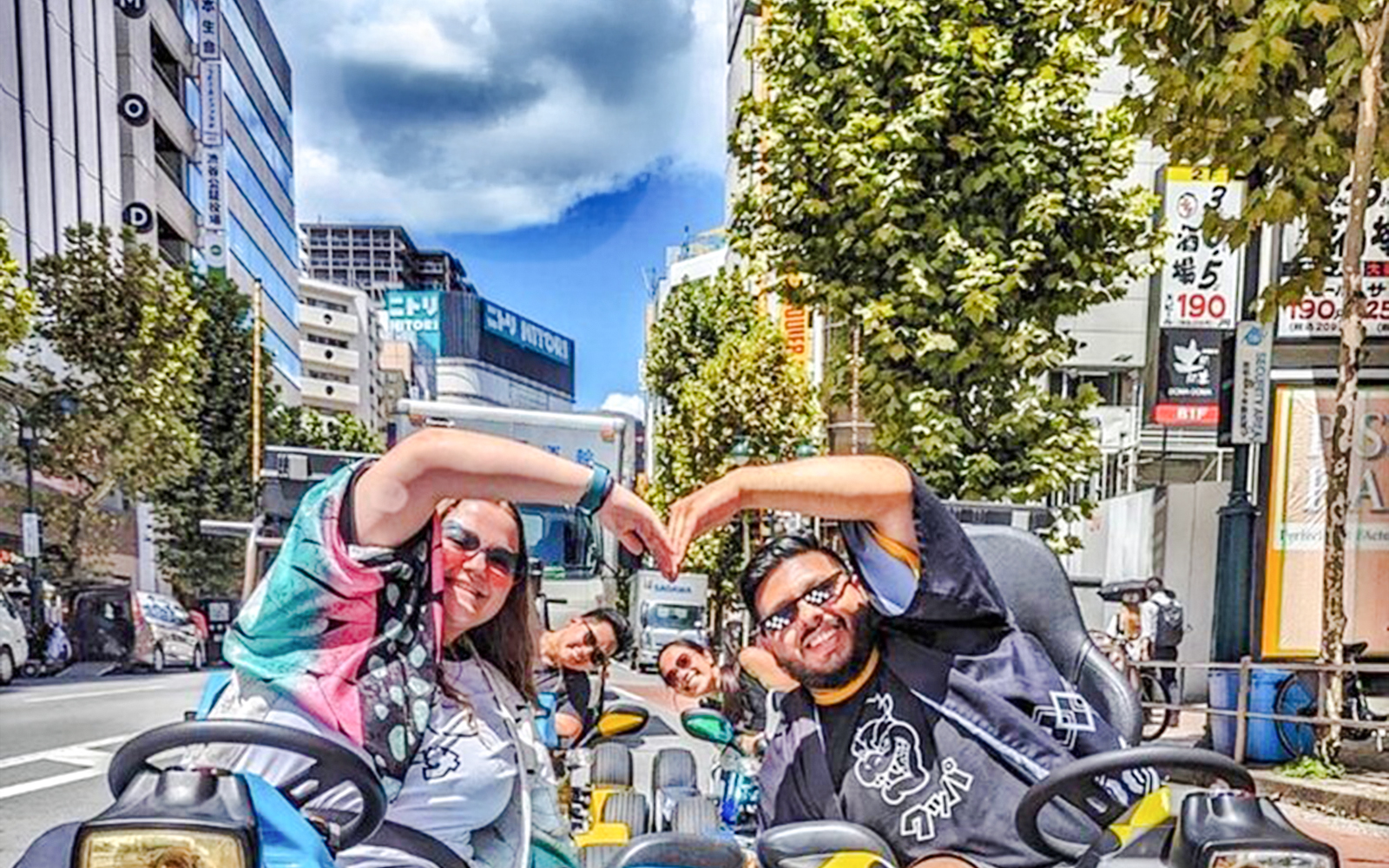 Two women in costume form heart shapes with hands while riding go-karts in Tokyo street.