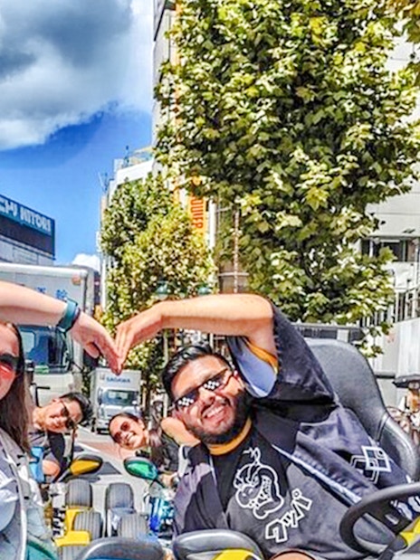 Two women in costume form heart shapes with hands while riding go-karts in Tokyo street.