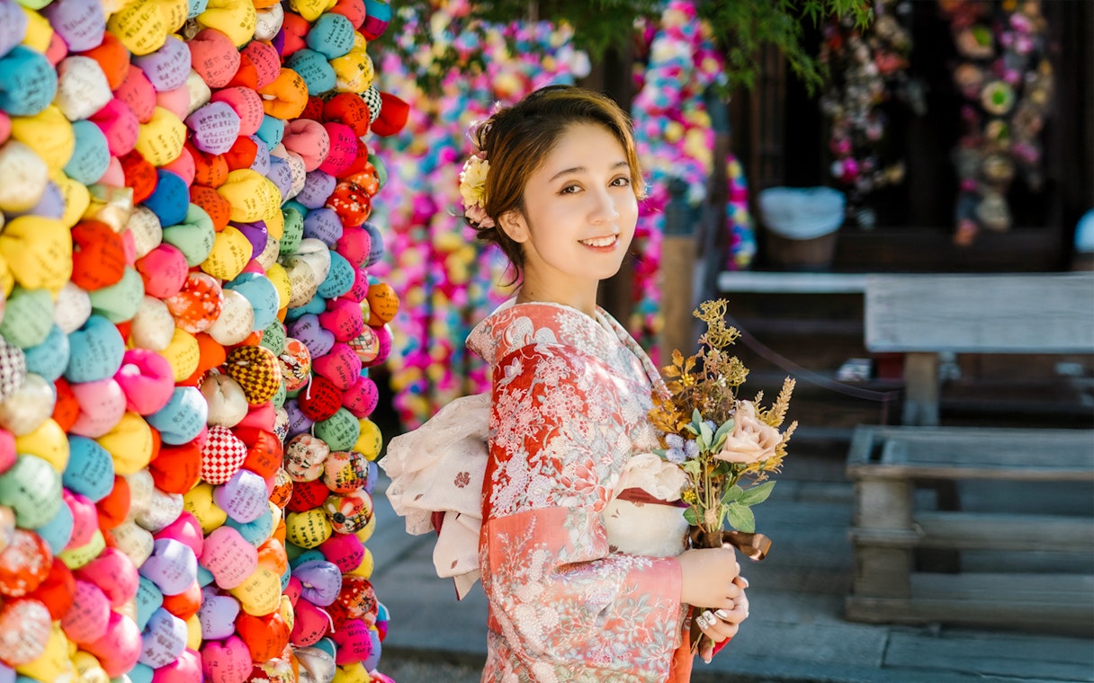 Person in traditional kimono holding flowers, standing by colorful prayer balls in Kyoto.