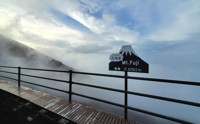 Mt. Fuji summit sign at 3,250 meters with misty clouds and wooden pathway.