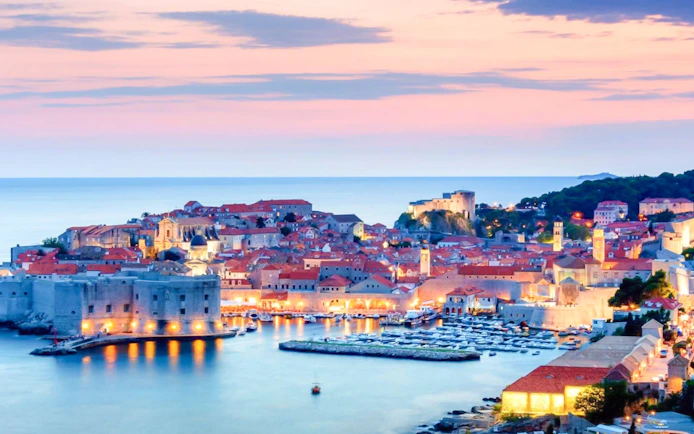 Dubrovnik Old Town at sunset with illuminated city walls and harbor.