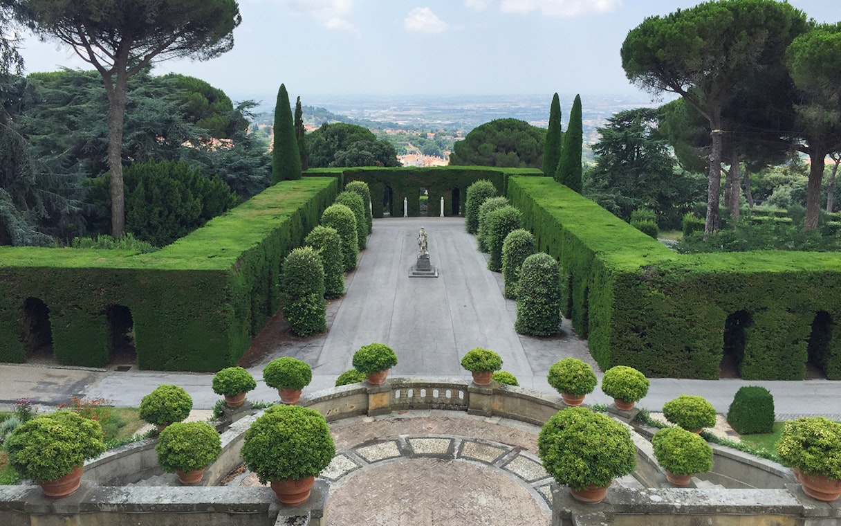Castle Gandolfo garden with manicured hedges and central statue, overlooking scenic landscape.