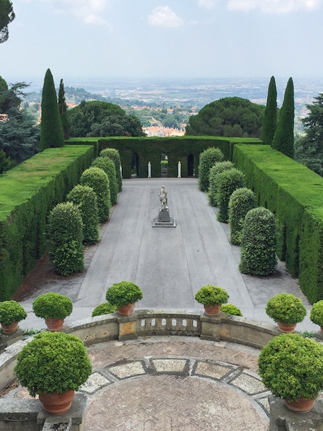 Castle Gandolfo garden with manicured hedges and central statue, overlooking scenic landscape.