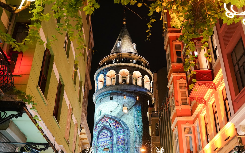 Galata Tower illuminated at night in Beyoglu, Istanbul, surrounded by colorful buildings.