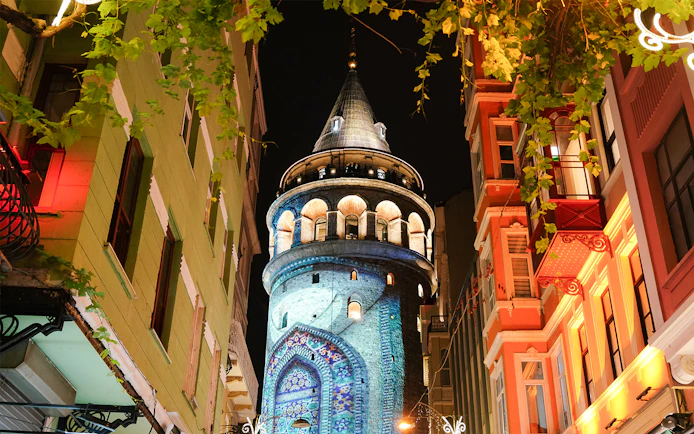Galata Tower illuminated at night in Beyoglu, Istanbul, surrounded by colorful buildings.
