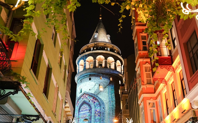 Galata Tower illuminated at night in Beyoglu, Istanbul, surrounded by colorful buildings.