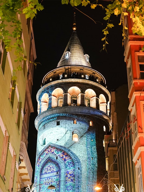 Galata Tower illuminated at night in Beyoglu, Istanbul, surrounded by colorful buildings.