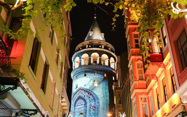 Galata Tower illuminated at night in Beyoglu, Istanbul, surrounded by colorful buildings.