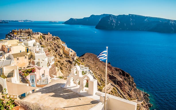Santorini coastline with Greek flag, white buildings, and view of Oia at sunset.