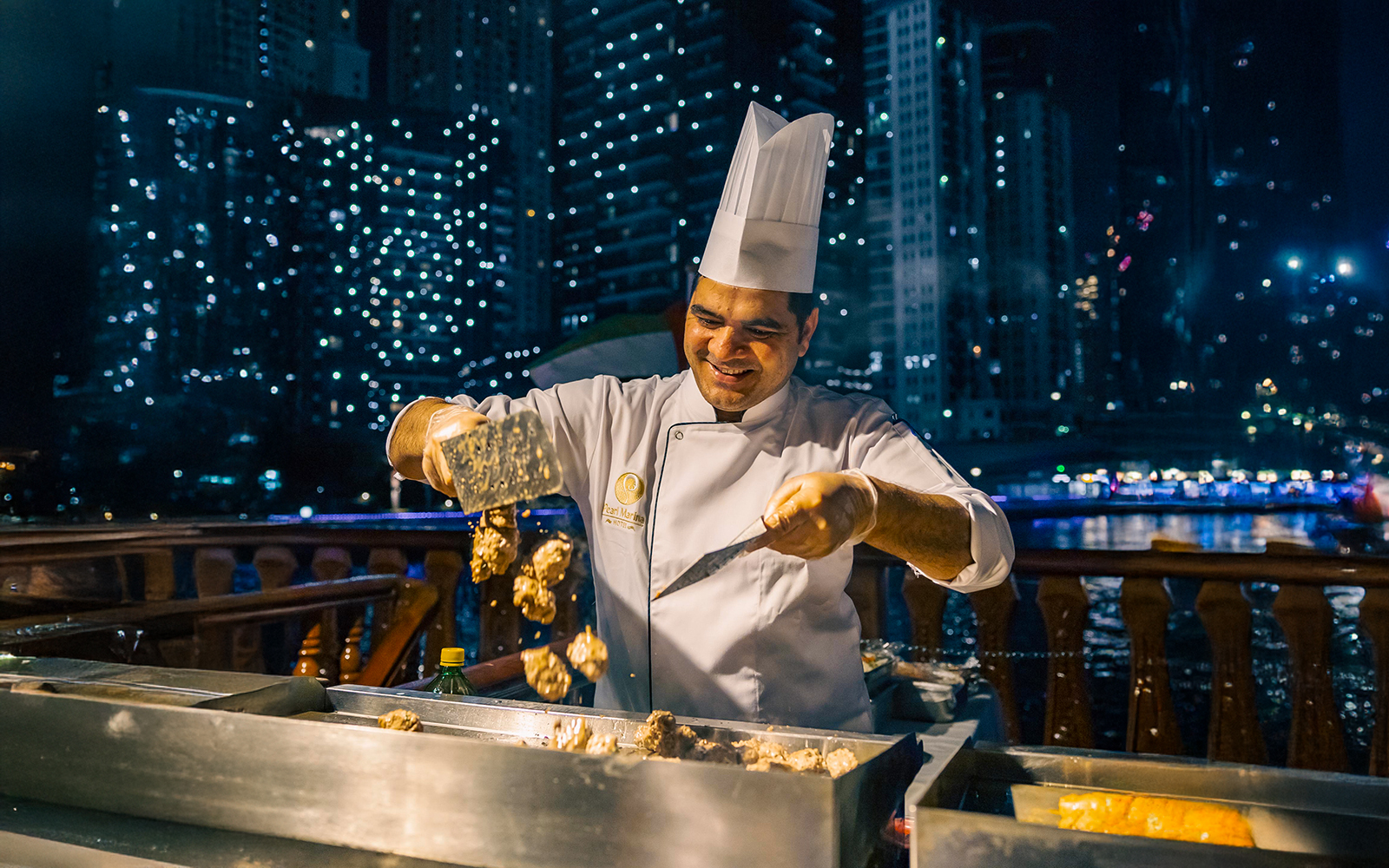 Chef preparing food on a Dubai Marina dhow cruise with city lights in the background.