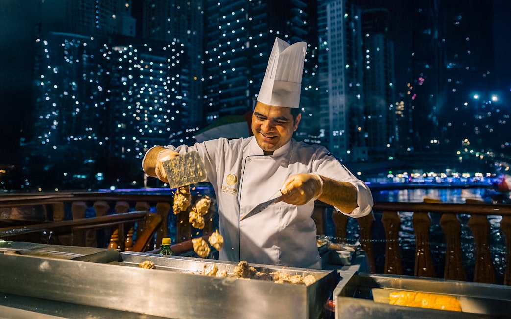 Chef preparing food on a Dubai Marina dhow cruise with city lights in the background.